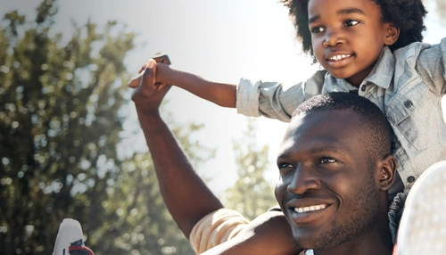 Foto de um homem sorrindo com uma criança sentada sob os seus ombros de braços erguidos para o ar, ao fundo da imagem aparece o céu e os galhos de árvores, com folhas. O homem é adulto, preto, de cabelo curto. A criança é preta, tem cabelos cacheados médio escuro, usa uma blusa social e calça jeans. Ambos olham para o horizonte num dia ensolarado. 